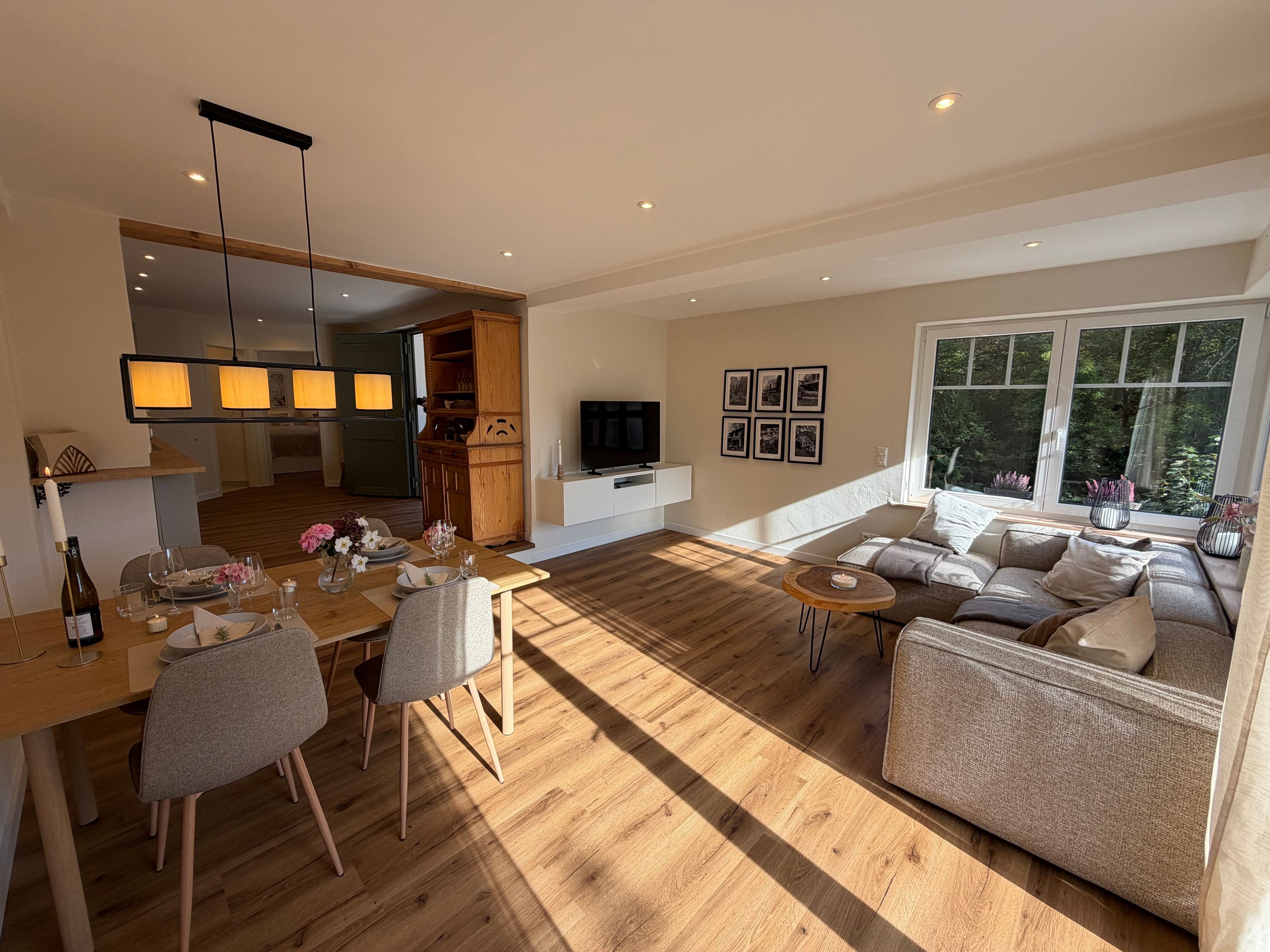 Een ruime woonkamer met een houten vloer, een eettafel voor twee personen, een bank en een televisie op een witte dressoir.