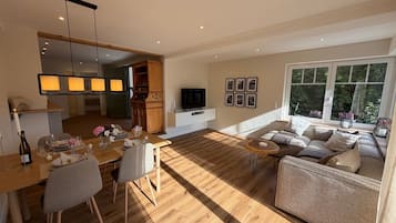 Een ruime woonkamer met een houten vloer, een eettafel voor twee personen, een bank en een televisie op een witte dressoir.