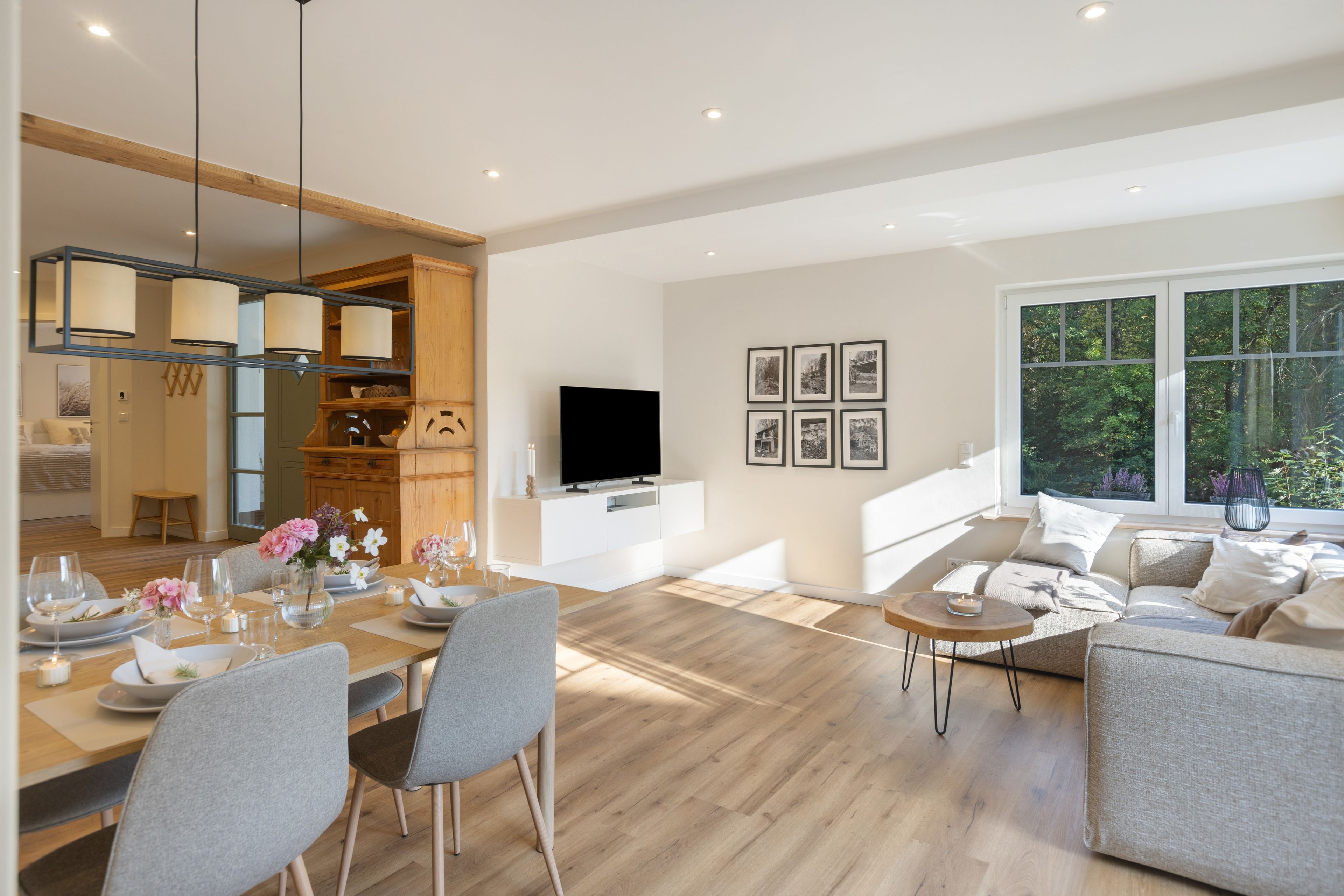 A modern living room with a dining area, a TV, and a large window with a view of greenery.