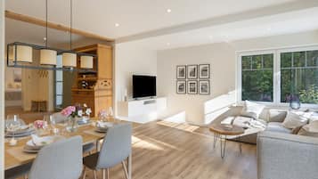 A modern living room with a dining area, a TV, and a large window with a view of greenery.