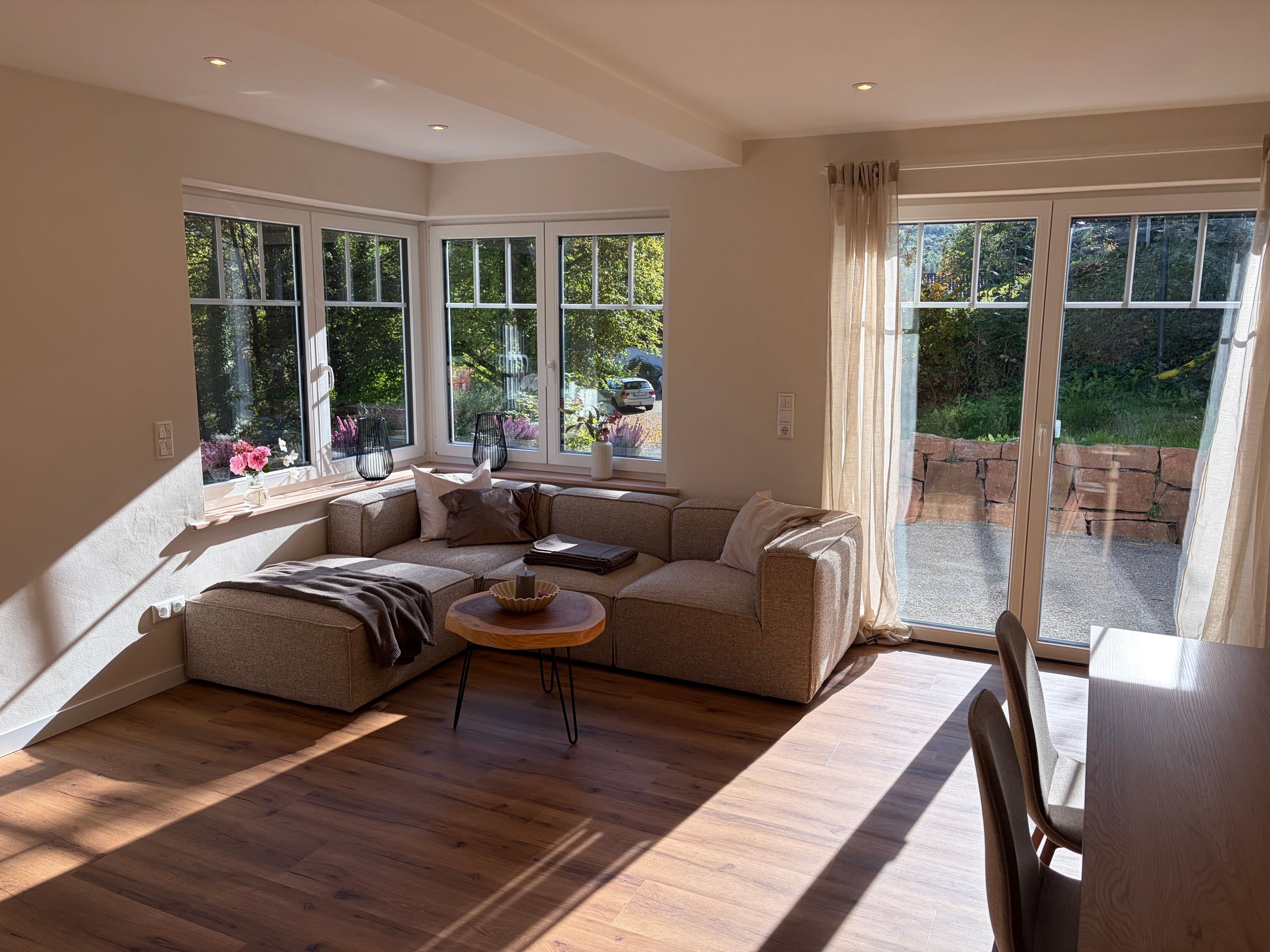 A modern living room with a sectional sofa, a round coffee table, and a dining table. Large windows provide a view of the garden.