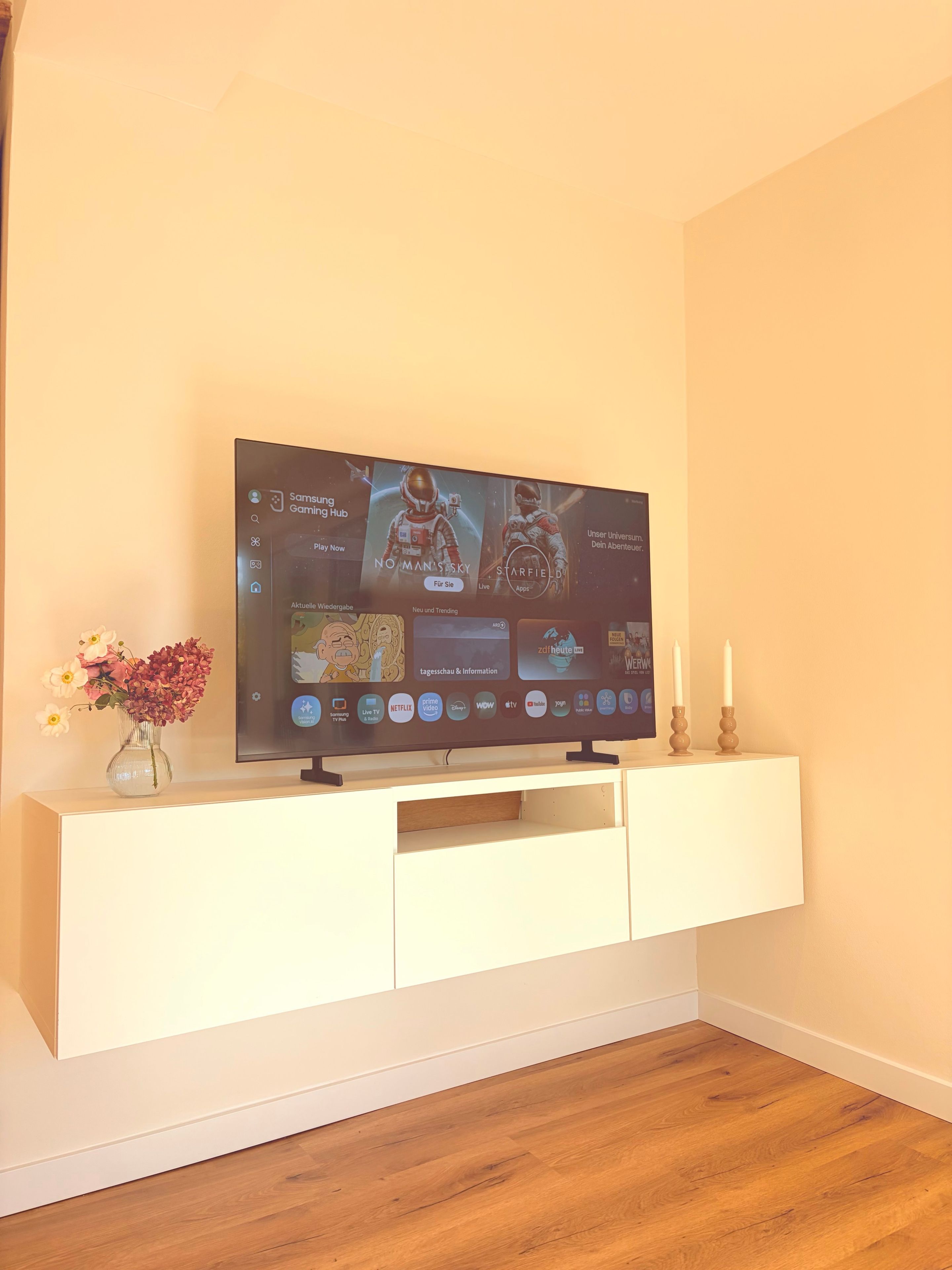 A flat-screen TV mounted on a white cabinet with a flower vase and candles on top.