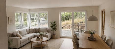 A spacious living area with a sofa, a dining table, and chairs, large windows with a view of the outdoors, and a ceiling light fixture.