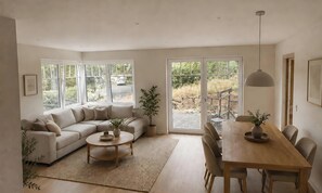 A spacious living area with a sofa, a dining table, and chairs, large windows with a view of the outdoors, and a ceiling light fixture.