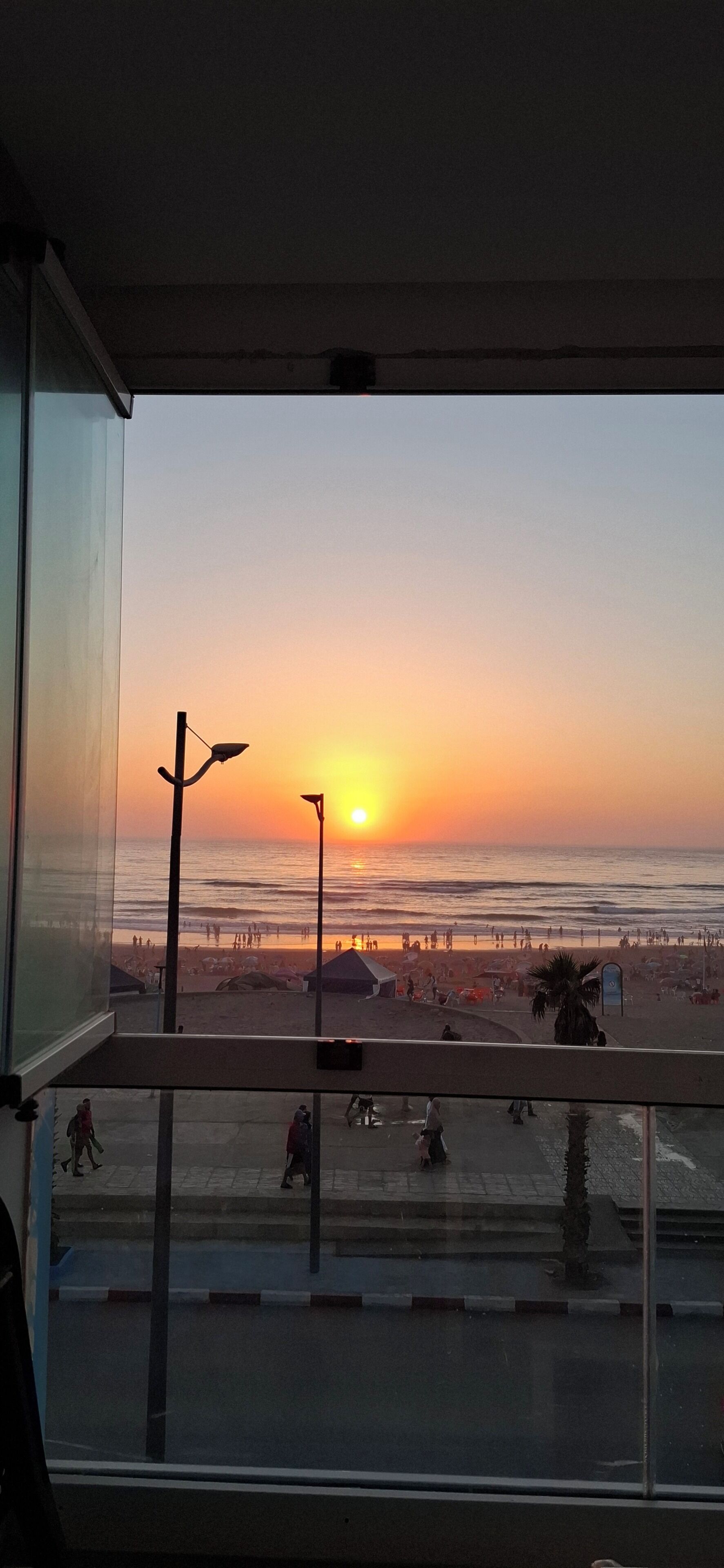 A sunset view from a balcony with a railing, a street below, and a beach with people and a pier in the distance.