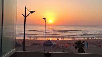 A sunset view from a balcony with a railing, a street below, and a beach with people and a pier in the distance.