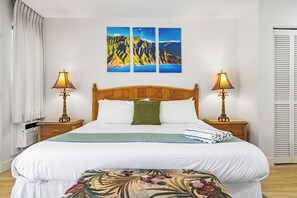 Una camera d'albergo con un letto in legno, due abat-jour, un cuscino decorativo verde e un pouf a motivi floreali.