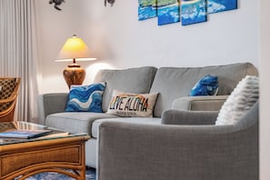 A living room with a grey sofa, two cushions, a lamp, a wicker chair, and a coffee table. - Maui Kamaole #K-104 Quiet Location, Close to Resort Pool, A/C (Kihei)