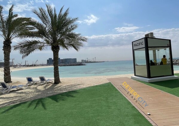 Une zone aménagée au bord de la plage, avec des palmiers, des chaises longues, un kiosque en verre portant l’inscription « PACIFIC » et un panneau indiquant « DESERT CITY STAYS ».