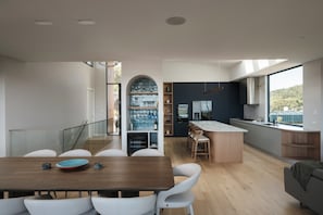 A modern kitchen with a central island, a dining area with a wooden table and white chairs, and a view of the outdoors through large windows. - Escape To The Bay – Grand Luxury with heated pool and spa (Nelson Bay)
