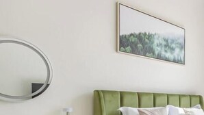 Makuuhuoneessa on vihreä, verhoiltu sängynpääty, valkoinen päiväpeite, pyöreä peili ja työpöytä tuolilla.