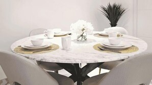 Uma mesa de jantar redonda com cadeiras brancas, um vaso de flores brancas e jogos americanos dourados.