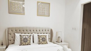 Quarto de hotel com cabeceira estofada, cama com roupa de cama branca e almofadas estampadas, duas obras de arte abstratas emolduradas na parede, mesa de cabeceira com abajur e criado-mudo com um pequeno vaso.