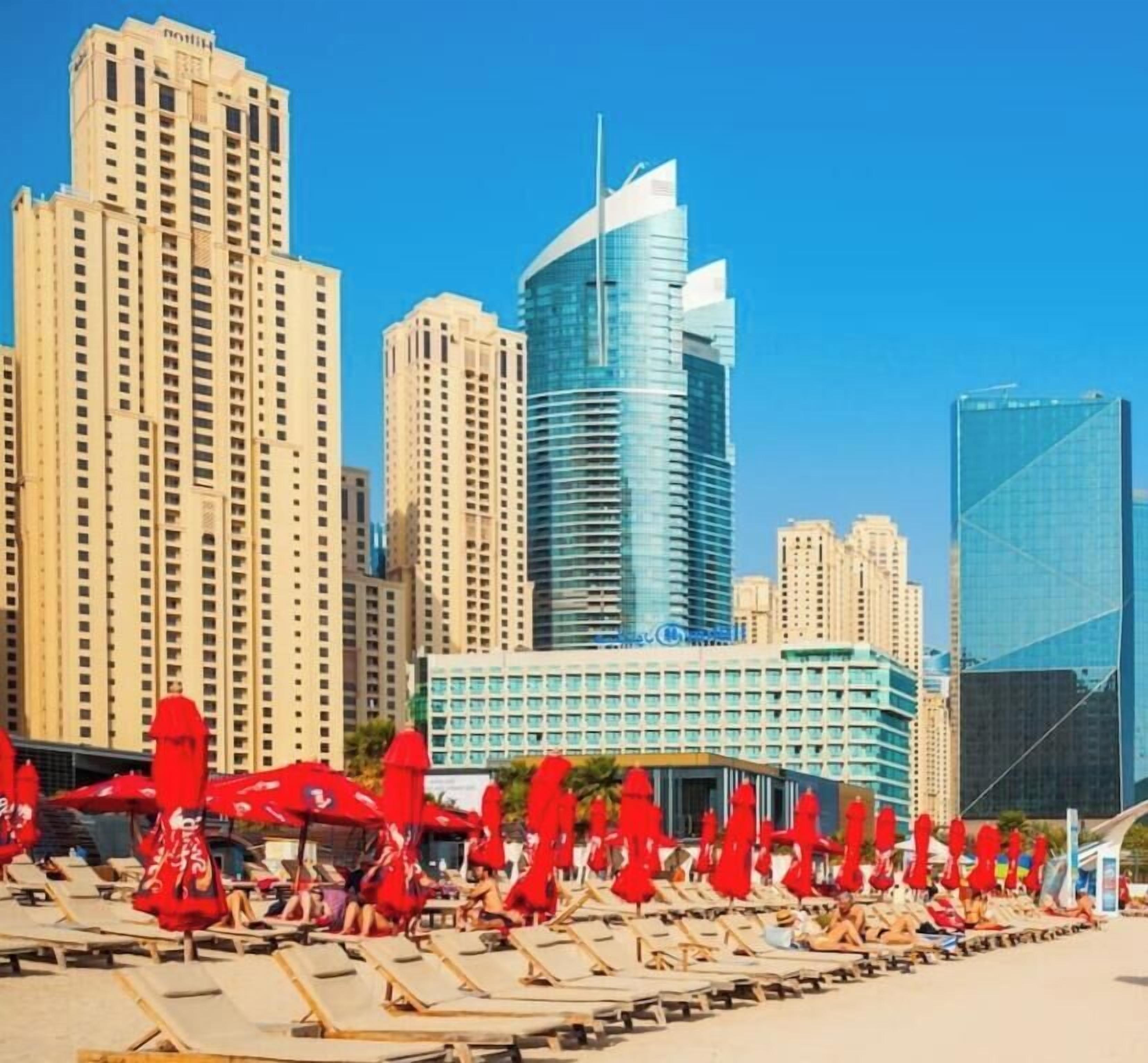 A beachfront hotel with multiple high-rise buildings in the background, a pool area with sun loungers, and a clear blue sky.