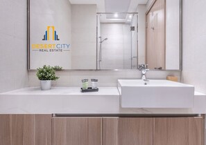 Um banheiro moderno com uma pia branca, um espelho e um chuveiro. Há um logotipo da Desert City Real Estate na parede.