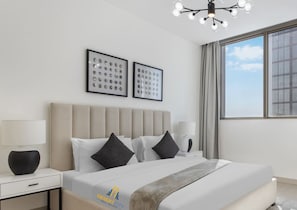 A modern hotel room with a large bed, a nightstand, a lamp, and a view of the sky through the window. - Peaceful Location | All Bills Included (Dubai)