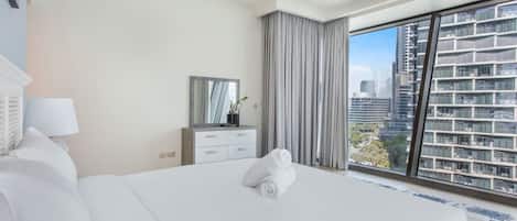 A hotel room with a large bed, a dresser, a mirror, and a view of the city through large windows.
