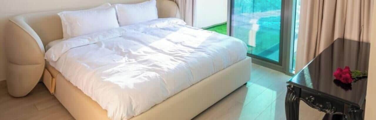 Un letto con copriletto e cuscini bianchi, un tavolino in vetro con un vaso e vista sul verde all'esterno.