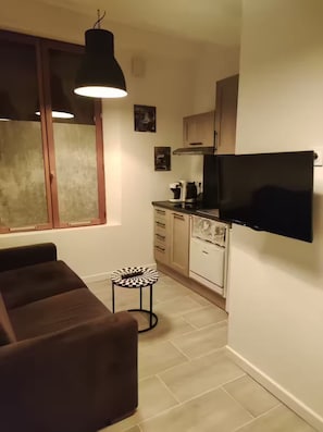 A compact living space with a sofa, a small round table, a television mounted on the wall, a kitchenette with a stove, and a window with blinds. - Small studio apartment right in the heart of downtown Meaux, with the train station less than a 10-minute walk away (Meaux)