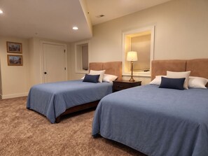 A hotel room with two beds, a nightstand, a lamp, a window with blinds, and a door.