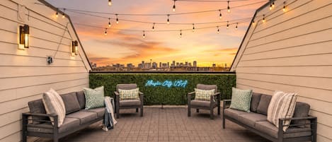 Terraza en la azotea con dos sofás, luces colgantes, un letrero de neón y vistas a la ciudad.