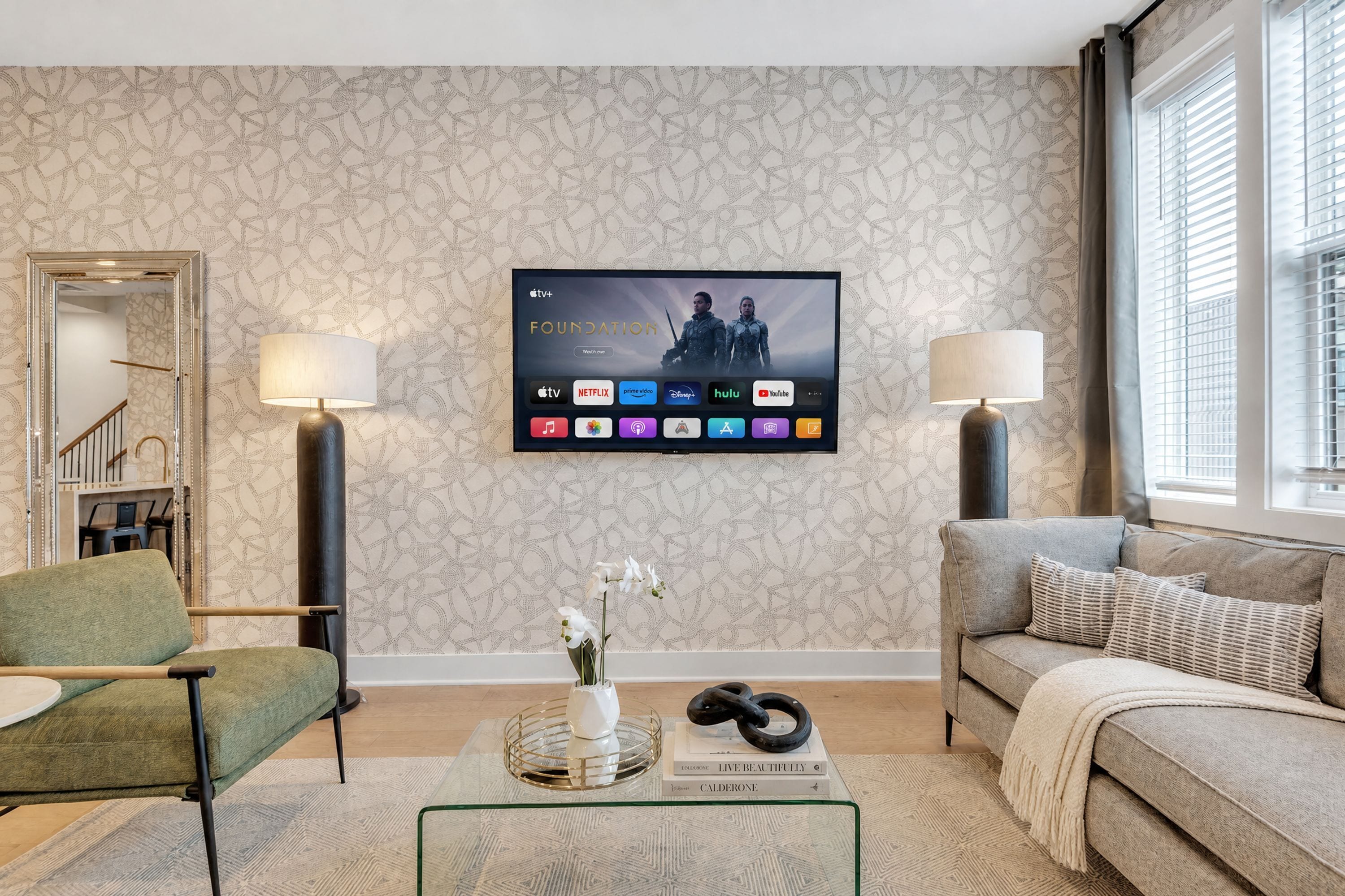 Una sala de estar con una televisión de pantalla plana montada en la pared mostrando la interfaz de un servicio de streaming, un sofá, una silla, una mesa de centro con objetos decorativos y un papel tapiz estampado.