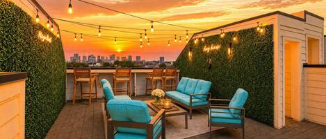 Un espacio al aire libre con sillas y una mesa, rodeado de vegetación y luces colgantes, con un horizonte urbano de fondo.
