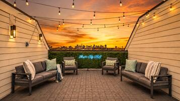 Un espacio exterior con dos sofás y sillones, luces colgantes y un letrero de neón que dice 'Music City USA' frente a un horizonte urbano al atardecer.