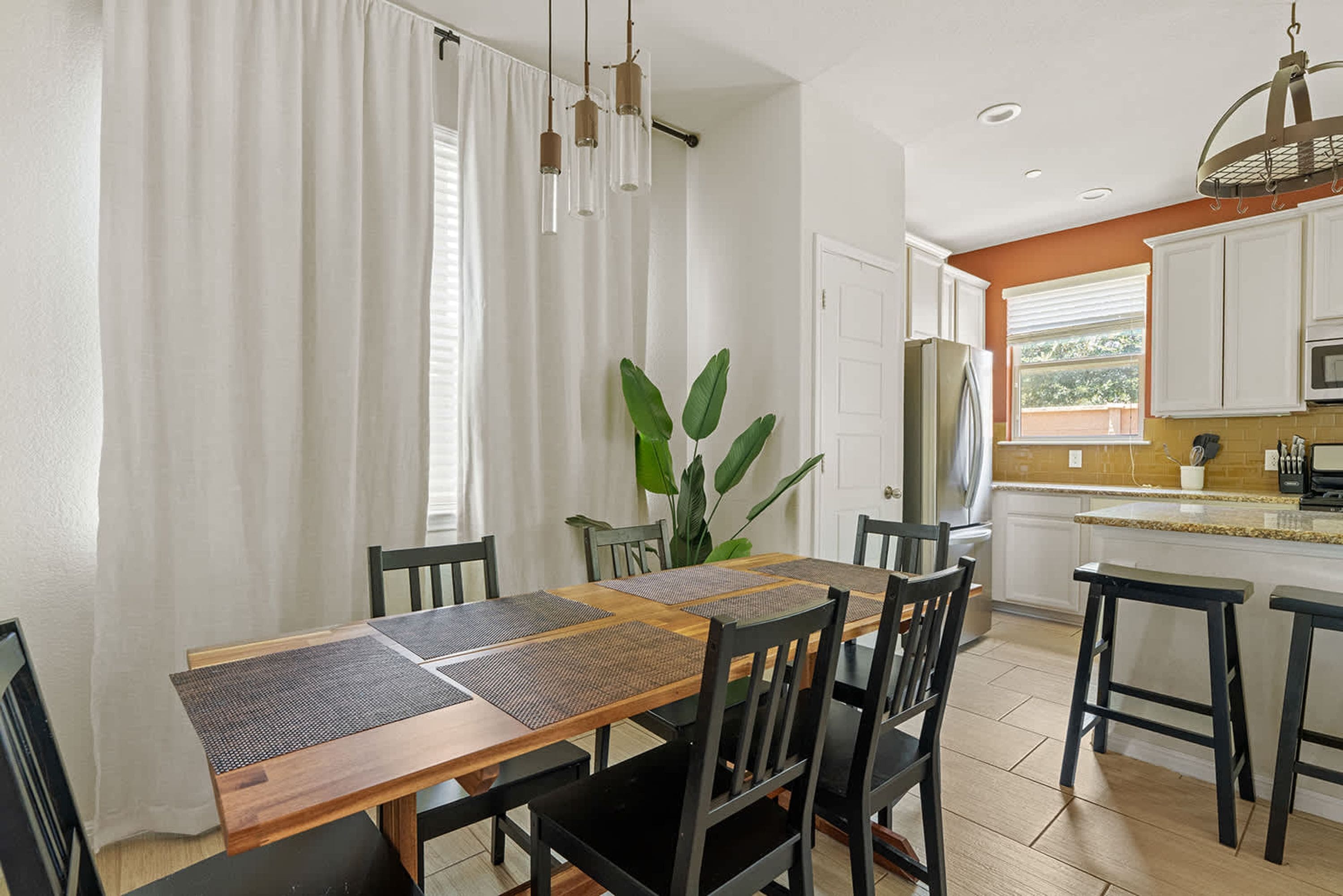 A dining area with a wooden table and chairs, a potted plant, and a kitchen with white cabinets and a window.
