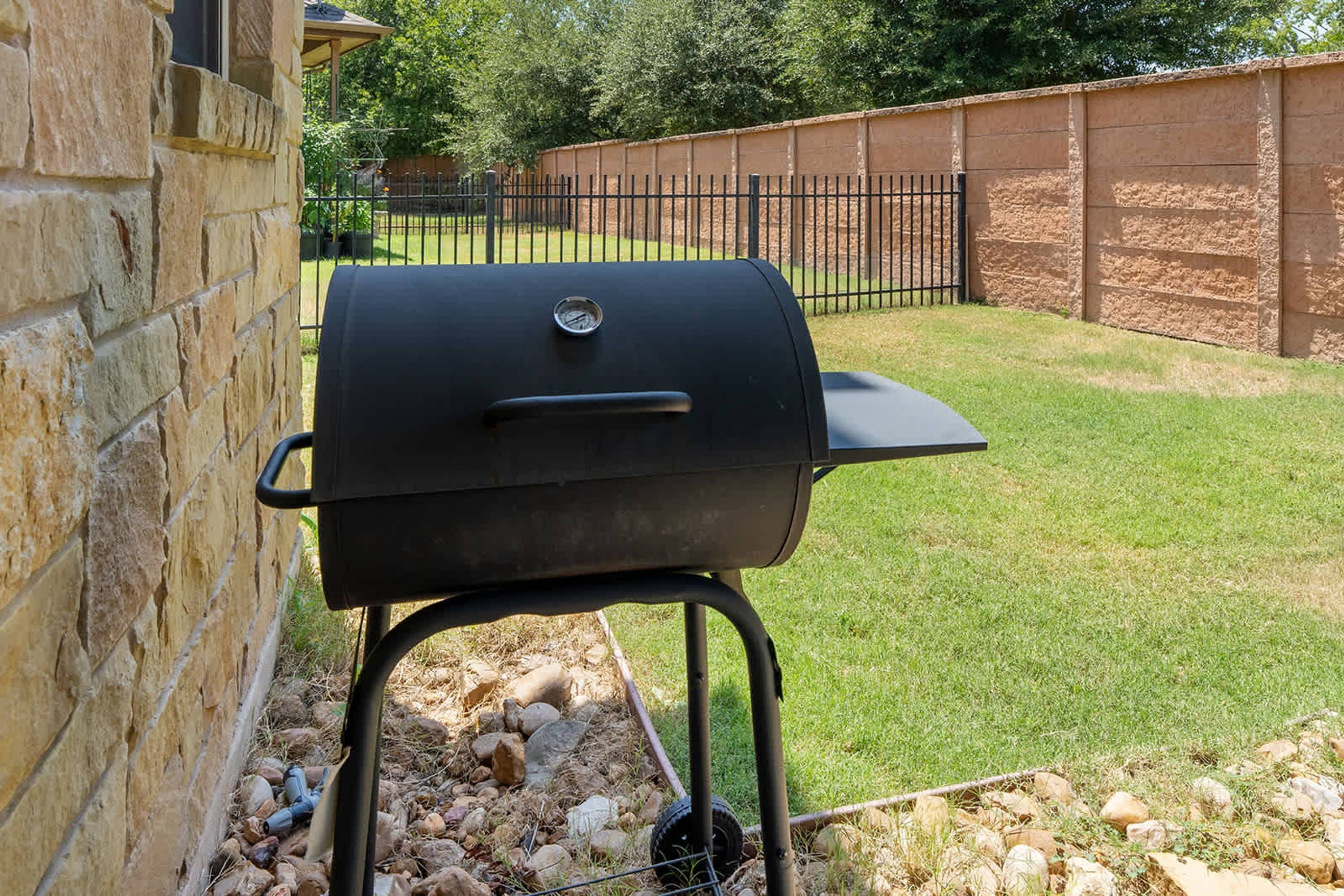 A barbecue grill positioned against a stone wall in a backyard with a grassy lawn and a metal fence in the background.