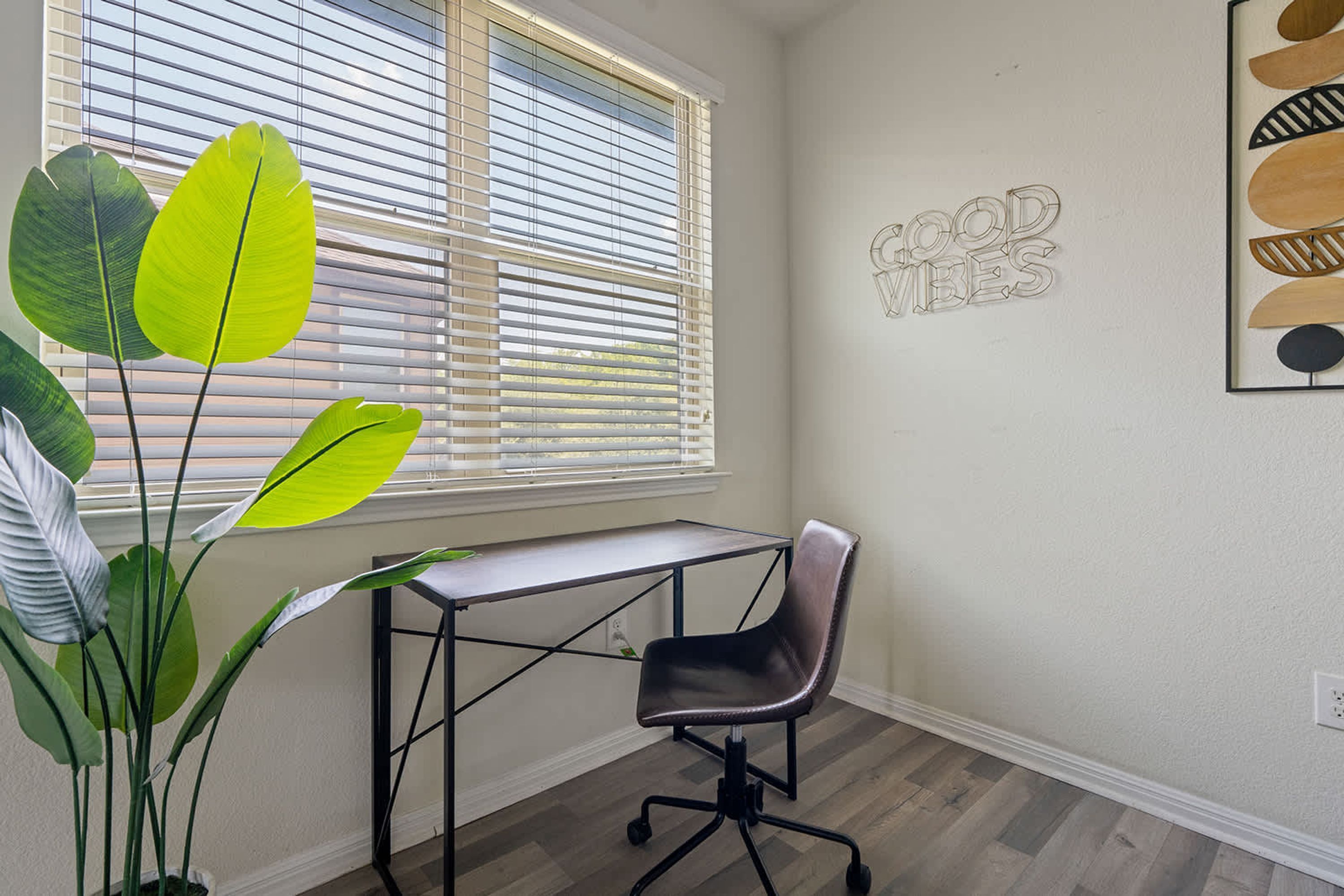 Una habitación con escritorio, silla, planta grande y ventana con persianas.