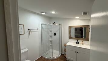 A bathroom with a glass-enclosed shower, a toilet, a sink with a mirror, and a framed picture on the wall.