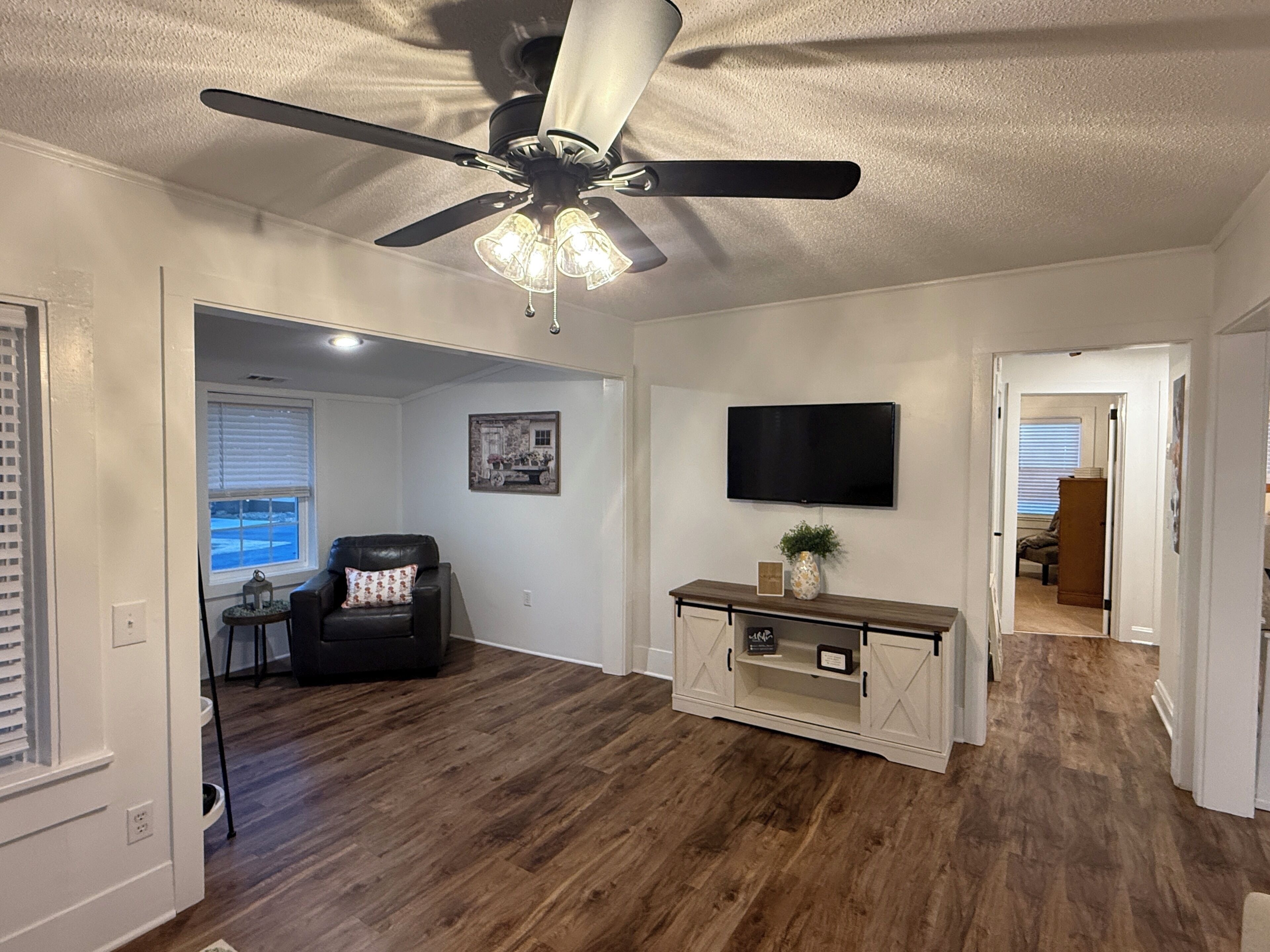A living room with a ceiling fan, a flat-screen TV mounted on the wall, and a wooden floor.