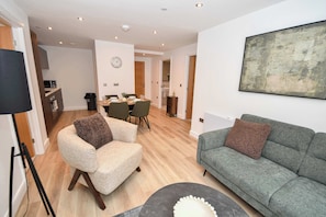 A modern living room with a grey sofa, a beige armchair, and a coffee table. There is a dining area with a table and chairs, and a kitchen in the background. - Bright 2 Bed Apartment in Central Leeds - Sleeps 4 (Leeds)