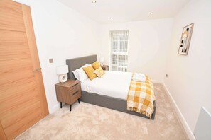 A bedroom with a bed, nightstand, lamp, window, and a door. - Bright 2 Bed Apartment in Central Leeds - Sleeps 4 (Leeds)