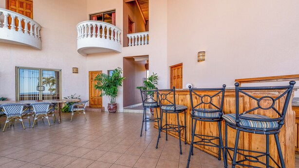 Um espaço amplo e interno com área de bar, com cadeiras de metal preto e balcão de madeira. Há várias portas e janelas, e uma grande planta em vaso é visível.
