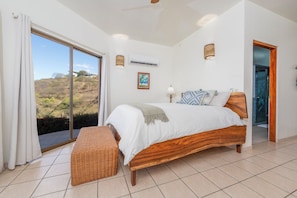 Un dormitorio con una cama grande, un banco de mimbre, vistas al exterior y una puerta al baño.