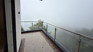 A balcony with a glass railing, a tiled floor, and a view of trees through foggy weather.
