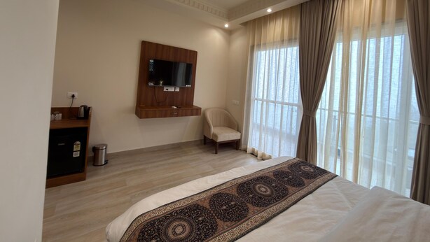 A hotel room with a large bed, a flat-screen TV mounted on the wall, a small seating area, and a window with curtains.