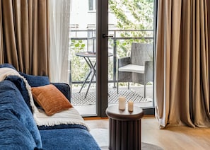 Um quarto com um sofá azul, uma mesa redonda pequena com velas, e vista para uma varanda externa com uma cadeira e mesa.