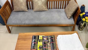 Un sofá de madera con cojines grises y almohadones decorativos, una mesa de centro de madera con un menú y una toalla blanca doblada, y un cuadro enmarcado sobre el sofá.