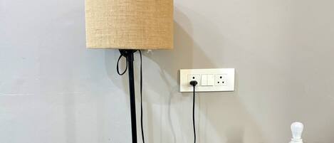 A floor lamp with a fabric shade, a power outlet, a light bulb, and a decorative vase with purple flowers on a wooden table.