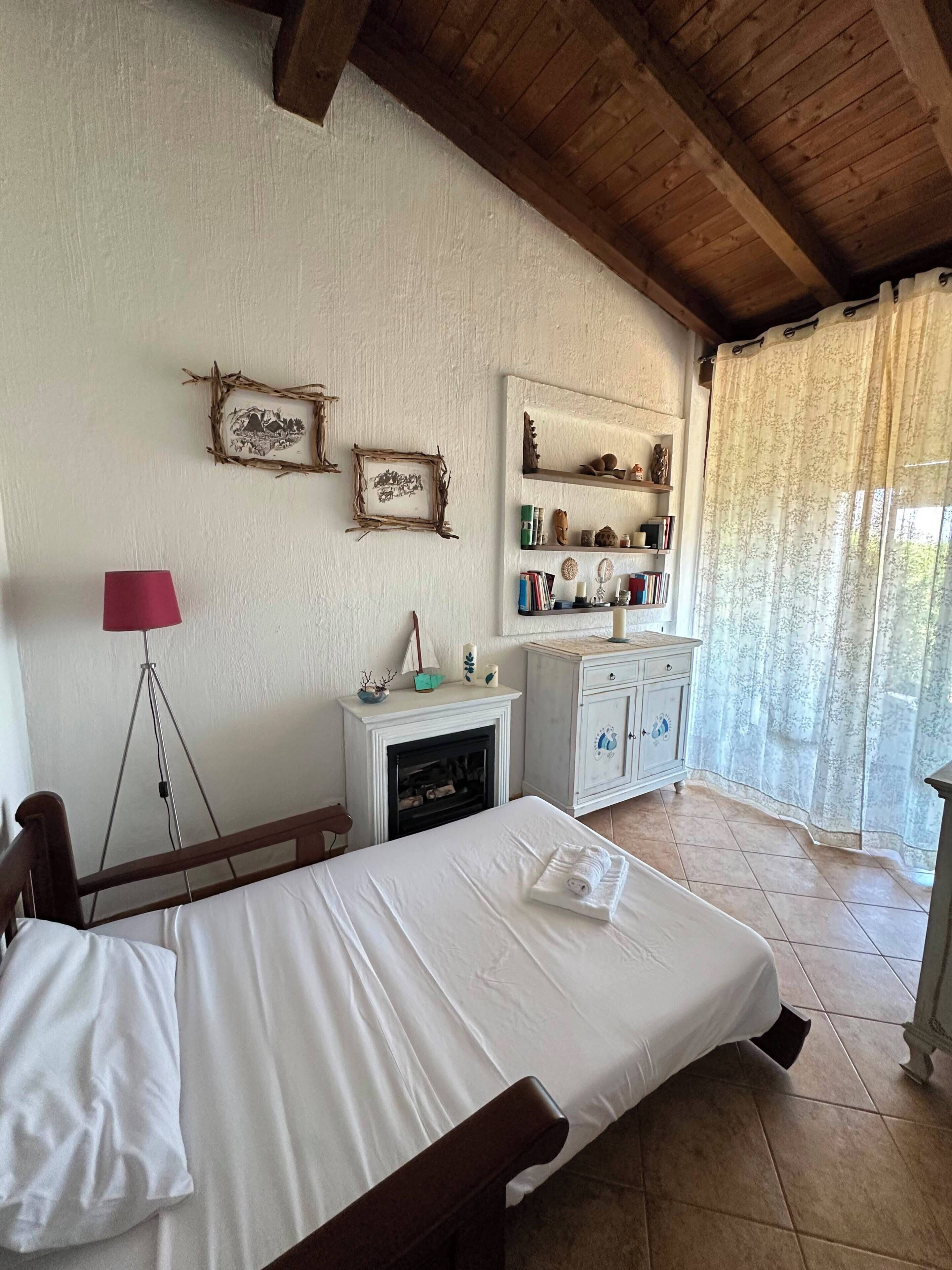A bedroom with a bed, a fireplace, a shelf with decorative items, and a window with curtains.
