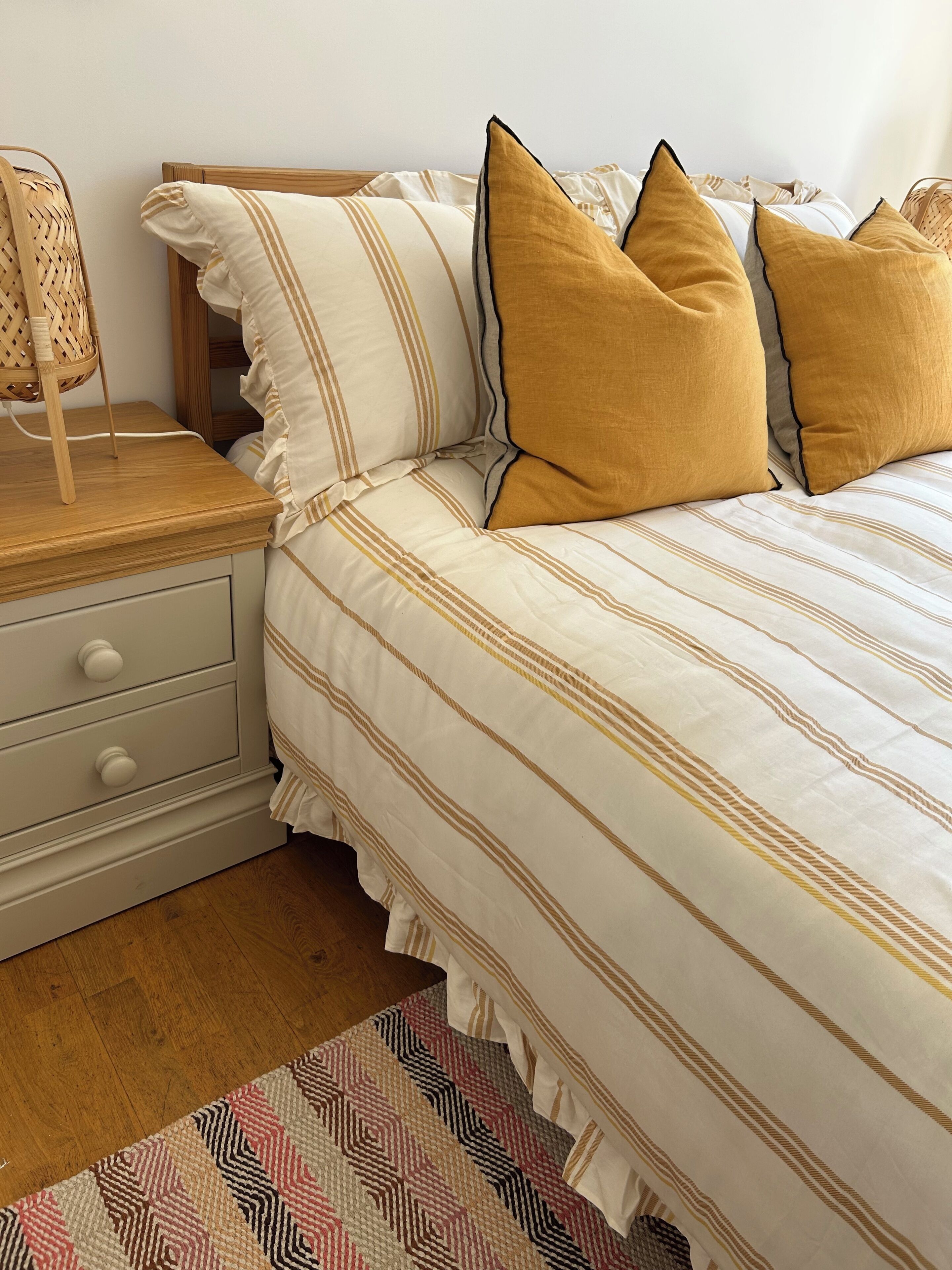 A neatly made bed with striped bedding and two pillows, a bedside table with a lamp, and a woven chair.