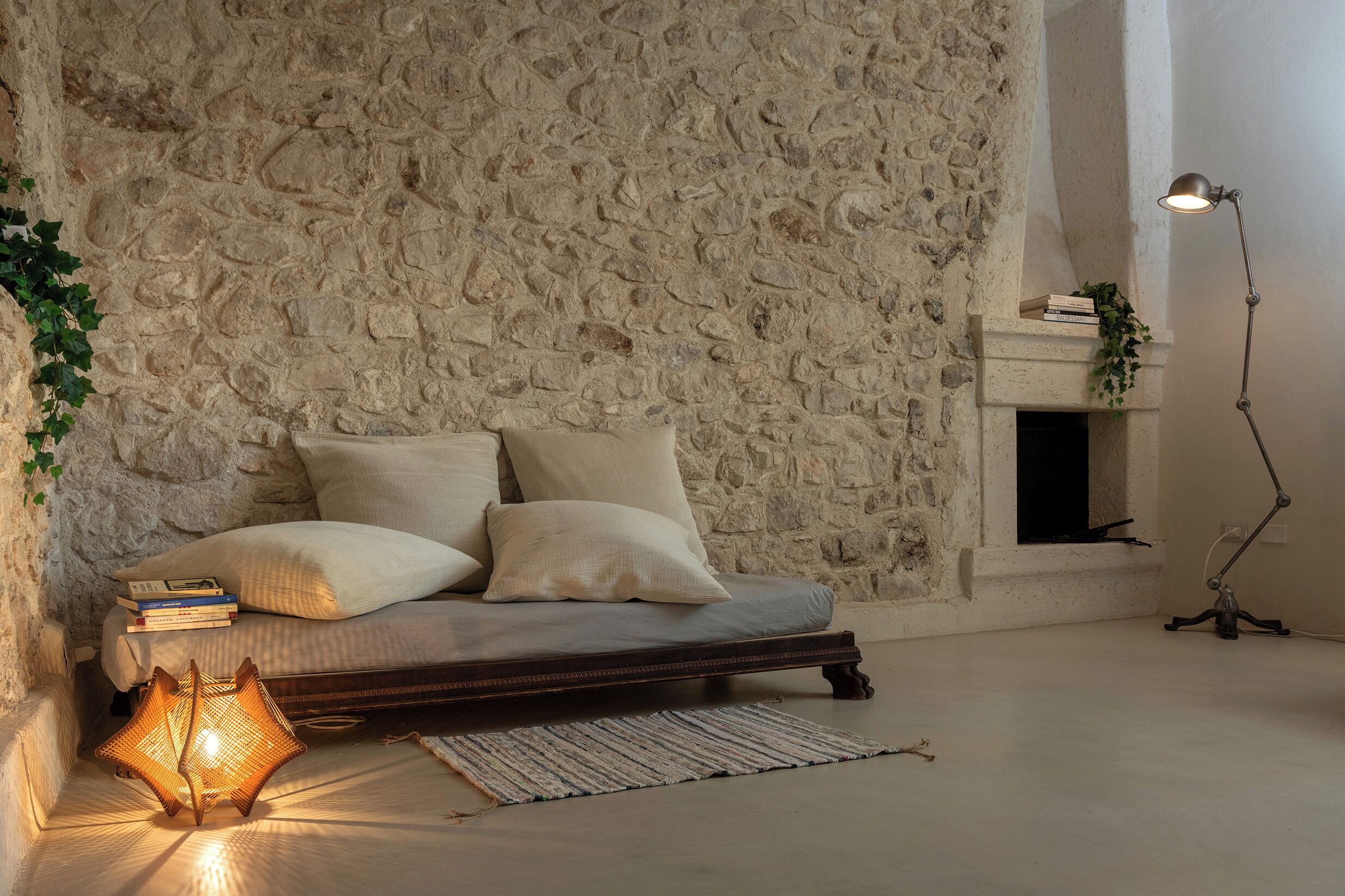A cozy room with a stone wall, a bed with pillows, a bedside table with books, a floor lamp, and a small plant.