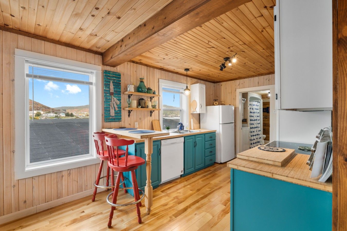 A kitchen with wooden walls, a wooden ceiling, a window with a view, a sink, a refrigerator, and a dining area with chairs.