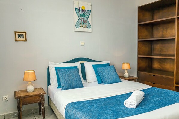 A neatly made bed with a blue blanket and pillows, two bedside lamps, a framed picture on the wall, and a wooden shelf unit.