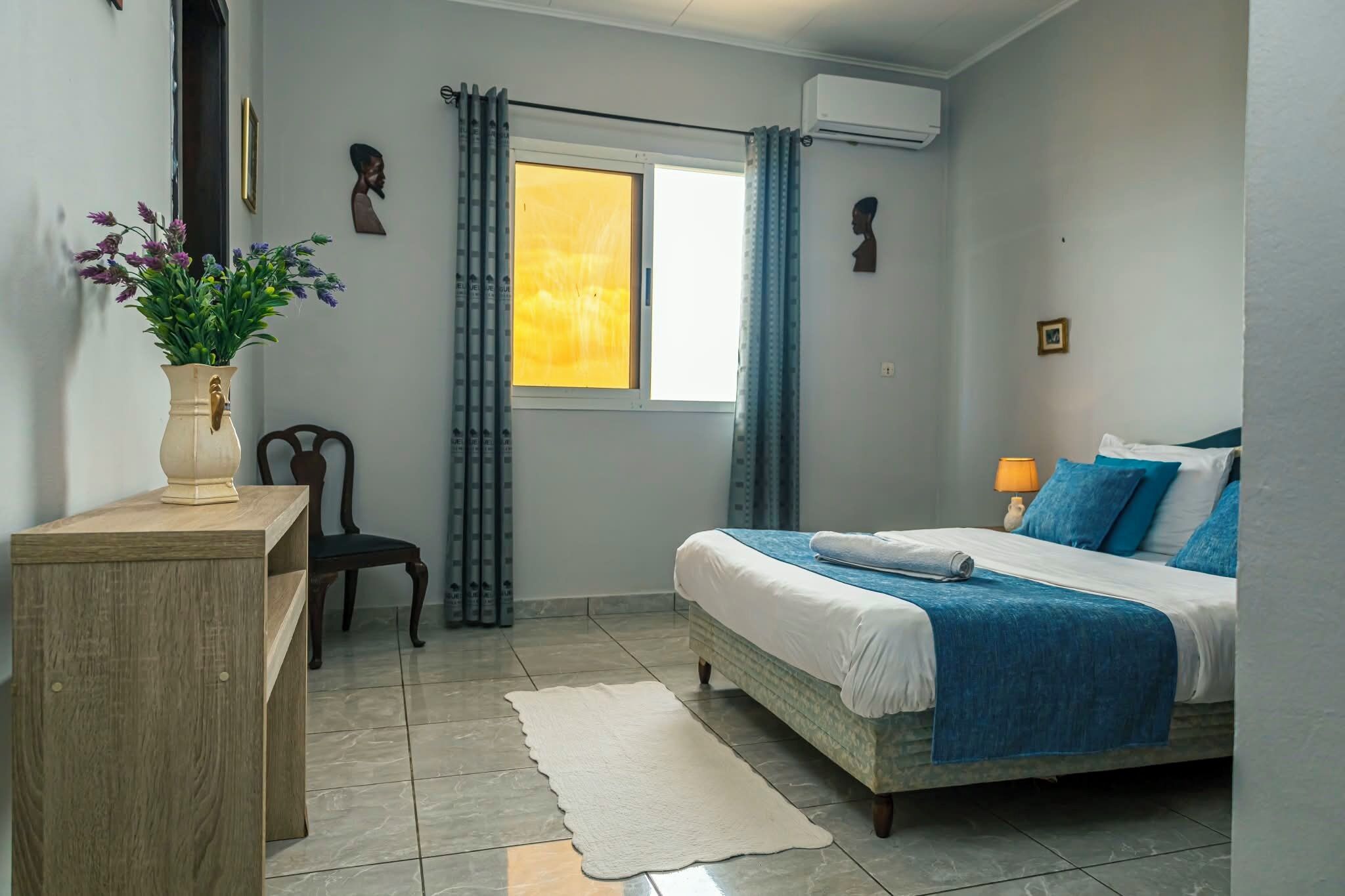 A bedroom with a bed, a chair, a desk, a window with curtains, and an air conditioning unit.