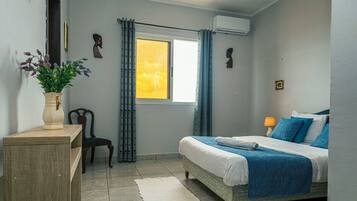 A bedroom with a bed, a chair, a desk, a window with curtains, and an air conditioning unit.