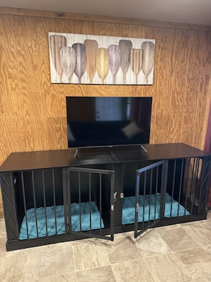 Una habitación con televisión montada en una pared de madera, un mueble de entretenimiento negro y una jaula para perro acolchada de color azul.